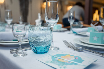 Festive table set with white cloth, plates, forks blue glasses and bomboniere for boy happy birthday party.