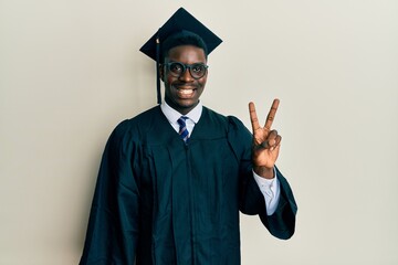 Handsome black man wearing graduation cap and ceremony robe showing and pointing up with fingers number two while smiling confident and happy.