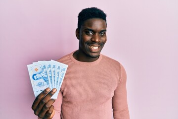 Handsome black man holding 50 thai baht banknotes looking positive and happy standing and smiling with a confident smile showing teeth