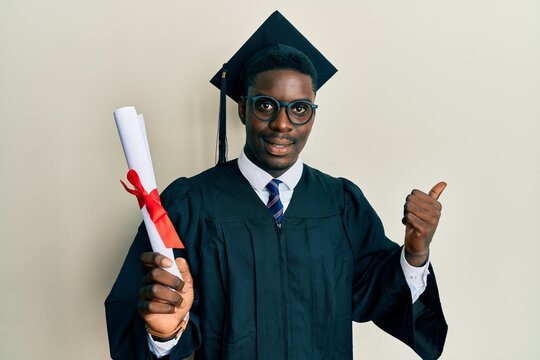 Handsome black man wearing graduation cap and ceremony robe holding diploma pointing thumb up to the side smiling happy with open mouth