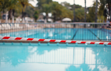 Red white tape in front of empty hotel resort swimming pool closed during evening cleaning, blurred water background