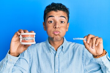 Young handsome african american man holding invisible aligner orthodontic and braces puffing cheeks with funny face. mouth inflated with air, catching air.