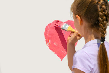 Baby girl draws a heart with a  brush on a white wall with pink paint