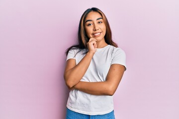 Young latin woman wearing casual white tshirt smiling looking confident at the camera with crossed arms and hand on chin. thinking positive.