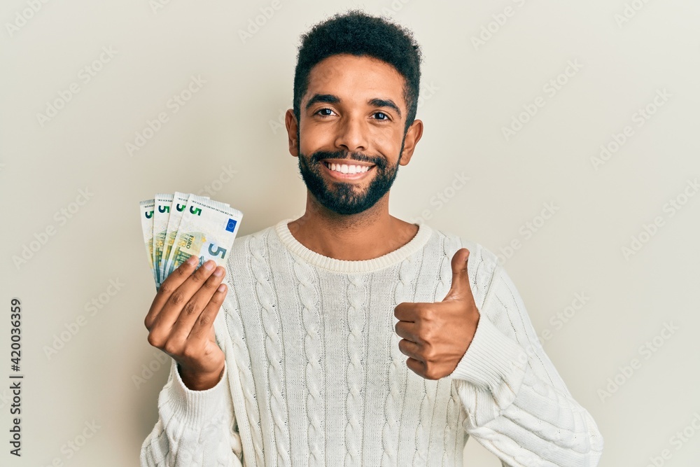 Wall mural Handsome hispanic man with beard holding bunch of 5 euro banknotes smiling happy and positive, thumb up doing excellent and approval sign