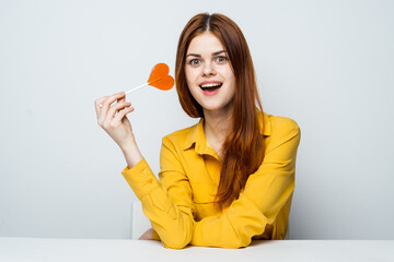 cheerful red-haired woman sits at the table in a yellow shirt with a heart-shaped lollipop