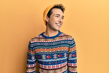 Young handsome man wearing wool hat and colorful sweater looking away to side with smile on face,...