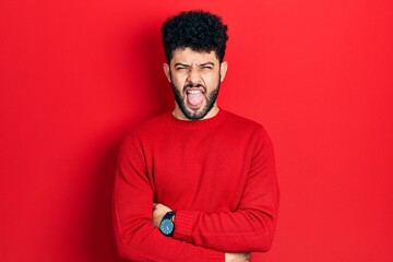 Young arab man with beard wearing casual red sweater sticking tongue out happy with funny expression. emotion concept.