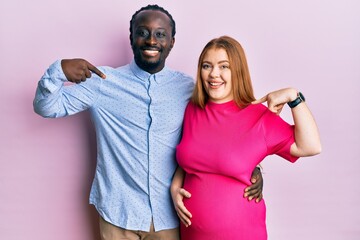 Obraz premium Young interracial couple expecting a baby, touching pregnant belly looking confident with smile on face, pointing oneself with fingers proud and happy.