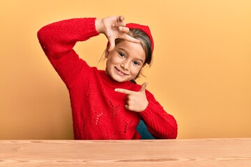 Little beautiful girl wearing casual clothes sitting on the table smiling making frame with hands and fingers with happy face. creativity and photography concept.