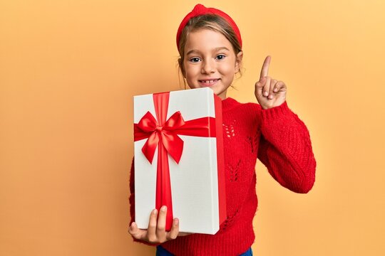 Little beautiful girl holding gift smiling with an idea or question pointing finger with happy face, number one