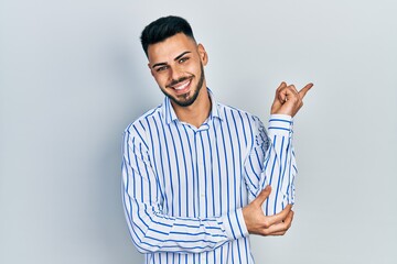 Fototapeta premium Young hispanic man with beard wearing casual striped shirt with a big smile on face, pointing with hand and finger to the side looking at the camera.