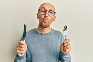 Bald man with beard holding fork and knife ready to eat looking at the camera blowing a kiss being...