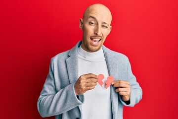 Bald man with beard holding broken heart paper shape winking looking at the camera with sexy...