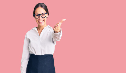 Beautiful brunette young woman wearing professional waitress apron smiling friendly offering handshake as greeting and welcoming. successful business.