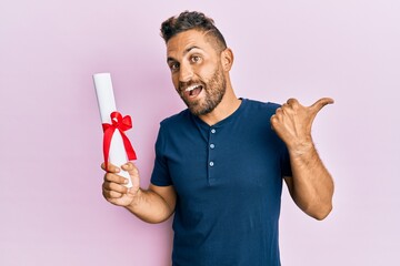Handsome man with beard holding graduate degree diploma pointing thumb up to the side smiling happy with open mouth - obrazy, fototapety, plakaty