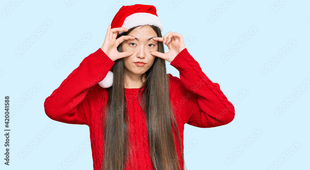 Sticker young chinese woman wearing christmas hat trying to open eyes with fingers, sleepy and tired for mor