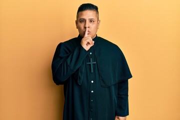 Young latin priest man standing over yellow background asking to be quiet with finger on lips....