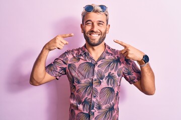 Handsome blond man on vacation wearing casual shirt and sunglasses over pink background smiling cheerful showing and pointing with fingers teeth and mouth. Dental health concept.