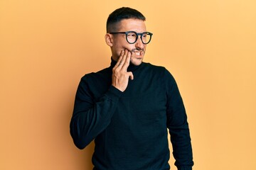 Fototapeta premium Handsome man with tattoos wearing turtleneck sweater and glasses touching mouth with hand with painful expression because of toothache or dental illness on teeth. dentist