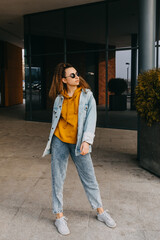 Young woman wearing denim clothes and a yellow sweatshirt, on city street buildings background.