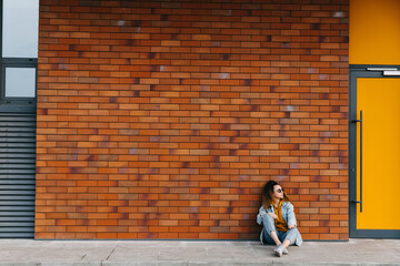 Young woman wearing 90s style denim clothes, sitting outdoors by a  brick wall.