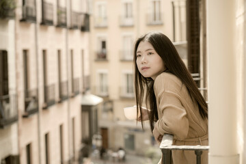 Asian woman in balcony thinking - outdoors lifestyle portrait of young beautiful and thoughtful Korean girl at drinking morning coffee looking to street indrawn and self-absorbed