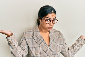 Young brunette woman wearing business jacket and glasses clueless and confused expression with arms...