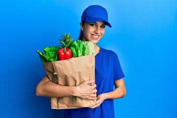 Young hispanic woman wearing courier uniform with groceries from supermarket looking positive and happy standing and smiling with a confident smile showing teeth