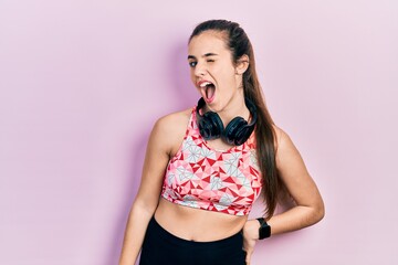 Young brunette teenager wearing gym clothes and using headphones winking looking at the camera with...