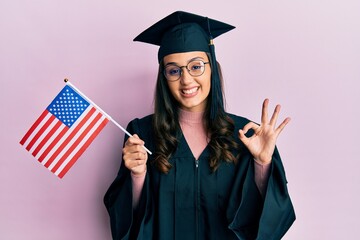 Young hispanic woman wearing graduation cap and ceremony robe holding usa flag doing ok sign with fingers, smiling friendly gesturing excellent symbol