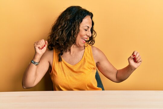 Beautiful middle age woman wearing casual clothes sitting on the table dancing happy and cheerful, smiling moving casual and confident listening to music