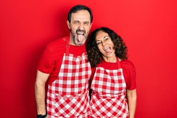 Middle age couple of hispanic woman and man wearing cook apron sticking tongue out happy with funny expression. emotion concept.