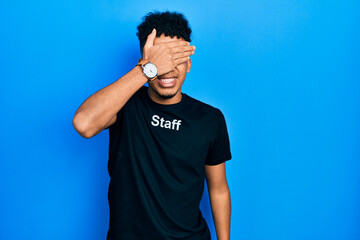 Young african american man wearing staff t shirt covering eyes with hand, looking serious and sad. sightless, hiding and rejection concept