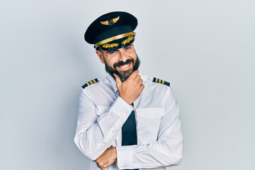 Young hispanic man wearing airplane pilot uniform looking confident at the camera smiling with crossed arms and hand raised on chin. thinking positive.
