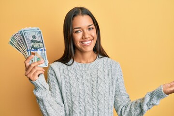 Beautiful hispanic woman holding dollars celebrating achievement with happy smile and winner expression with raised hand