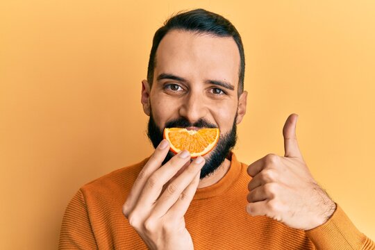 Young Man With Beard Holding Orange Slice On Mouth As Funny Smile Smiling Happy And Positive, Thumb Up Doing Excellent And Approval Sign