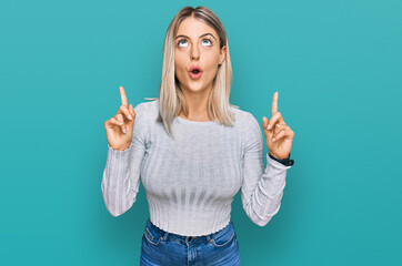 Beautiful blonde woman wearing casual clothes amazed and surprised looking up and pointing with fingers and raised arms.