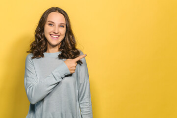 Happy and smiling young woman points away with a finger on a copy space, presenting promo or sale, cheerful girl wearing blue pullover showing on the novelty, empty space, mockup, isolated on yellow