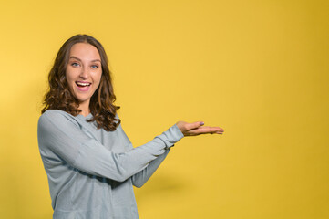 Fototapeta na wymiar Cheerful and happy young woman in casual wear points with hands to the empty space, excited curly lady happy to presenting novelty, copy space, mockup, isolated on the yellow