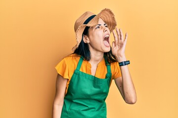 Beautiful young woman wearing gardener apron and straw hat shouting and screaming loud to side with hand on mouth. communication concept.