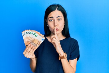 Beautiful young woman holding bunch of 50 euro banknotes asking to be quiet with finger on lips. silence and secret concept.