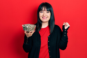 Young hispanic woman holding sunflower seeds bowl screaming proud, celebrating victory and success very excited with raised arm