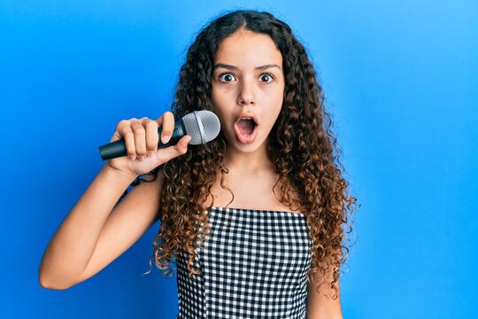Teenager hispanic girl singing song using microphone scared and amazed with open mouth for surprise, disbelief face