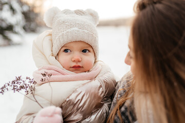 Young mom holding small beautiful baby girl in winter hat and with flower in her arm, outside, winter landscape