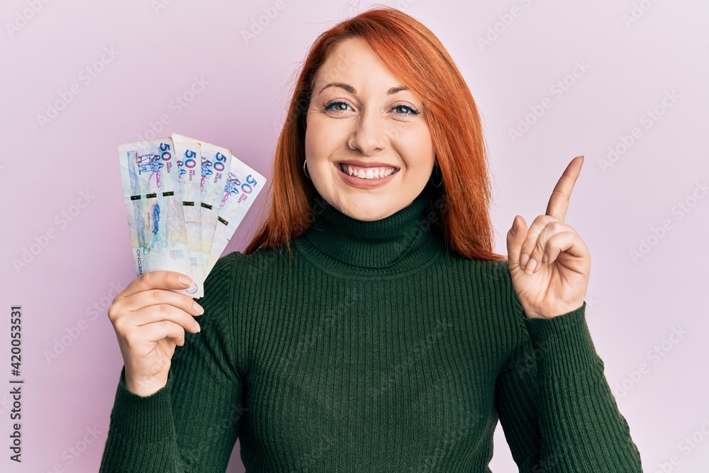 Poster beautiful redhead woman holding 50 colombian pesos banknotes smiling happy pointing with hand and fi