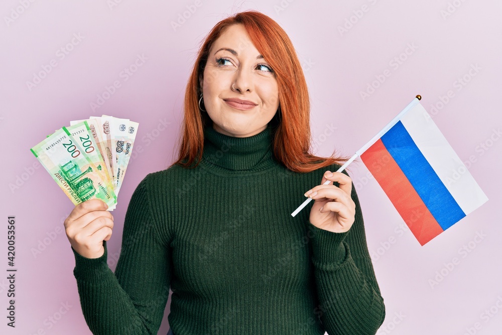 Canvas Prints Beautiful redhead woman holding russian ruble banknotes and russia flag smiling looking to the side and staring away thinking.