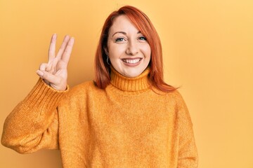 Beautiful redhead woman wearing casual winter sweater over yellow background showing and pointing up with fingers number three while smiling confident and happy.