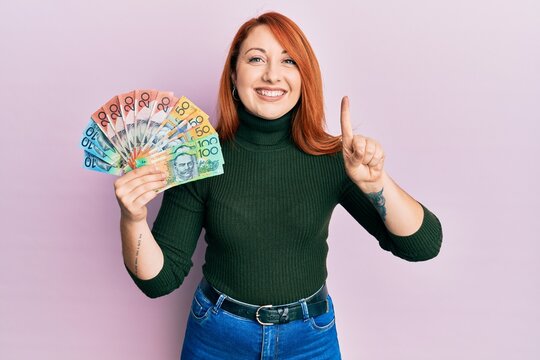 Beautiful redhead woman holding australian dollars smiling with an idea or question pointing finger with happy face, number one