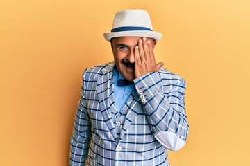 Mature middle east man with mustache wearing vintage and elegant fashion style covering one eye with hand, confident smile on face and surprise emotion.